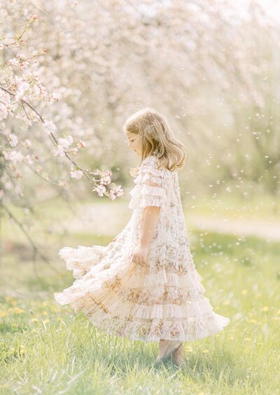 Young girl wearing a floral, ruffled dress next to a blooming tree with white flowers by Kansas City family photographer Jeanna Hayes
