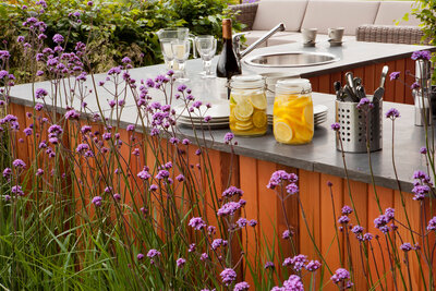 A chic outdoor kitchenette with a wooden countertop framed by tall, slender purple flowers, set against a backdrop of lush foliage.