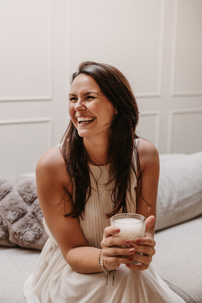 Mooie lachende vrouw met een latte machiatto in haar handen. Heeft een licht beige jurk aan. Een lichte achtergrond.
