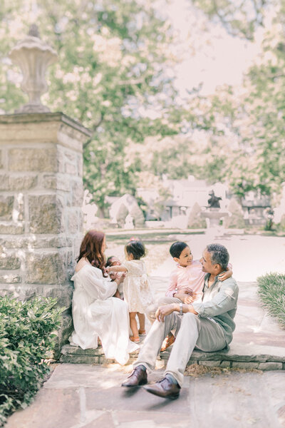 A family of five sits in a beautiful Kansas City garden. While mom holds her newborn, big sis looks on, while big brothers sits on his dad's lap who is lovingly smiling at him. 
