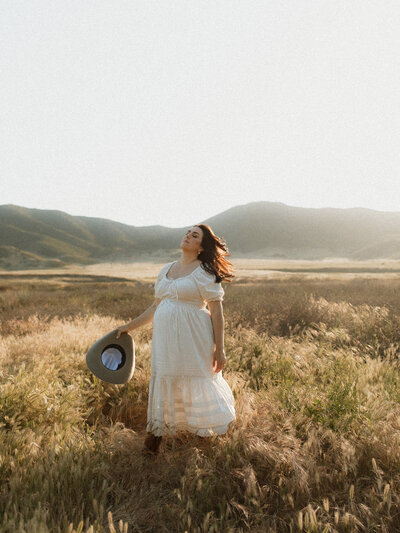 A pregnant woman in a white dress stands in a sun-drenched field, eyes closed and wind in her hair, holding a wide-brimmed hat at her side during golden hour in Temecula.
