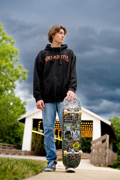 senior wearing a band sweatshirt and holding his skateboard looks off to his left with dark clouds all around him