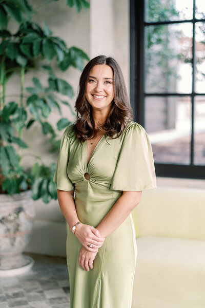 Headshot of Sarah in a light purple dress sitting in a white chair in front of a floral wall