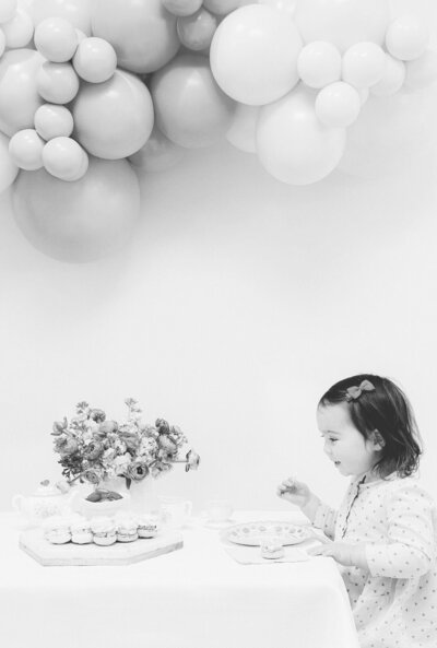 Black and white photo of a little girl at a table with flowers and cake underneath a balloon arch