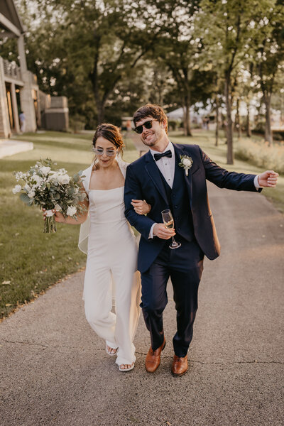 Bride and groom walk arm in arm down a path as they bump hips and look like they are having the time of their lives. The bride is rocking a white pantsuit and holding her bouquet in one arm while the other arm is linked with her new husband.