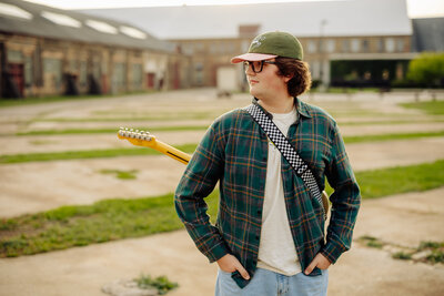 A person in a green cap, glasses, and checkered shirt stands outdoors in a courtyard with buildings and greenery, holding a guitar. Capturing life's melodies like this is the specialty of any Minnesota photographer skilled in Brainerd Lakes photography.