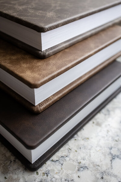 Stack of handcrafted leather-bound wedding albums in warm earth tones, displayed on a stone countertop.
