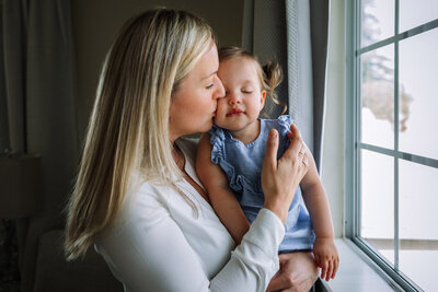 mom standing by window holding toddler girl and kissing her cheek as girl closes her eyes and leans in towards mom