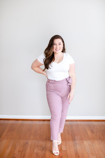 Woman posing for camera standing on hardwood floor in white shirt and purple pants, with grey background behind