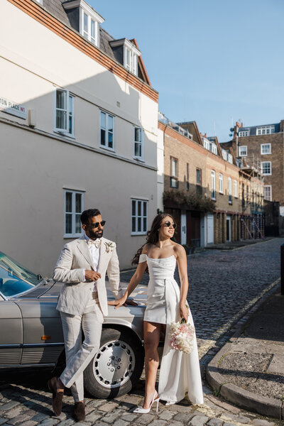 bride and groom standing next to convertible car in the sunshine on cobbled street