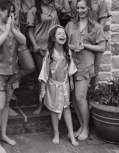 A joyful young girl in a satin robe and flower crown stands laughing among women in matching pajamas, exuding happiness in a relaxed setting.