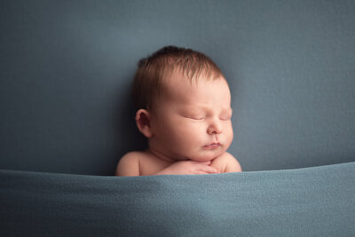 Newborn baby boy tucked in under a blue blanket sleeping with his face turned to his left.