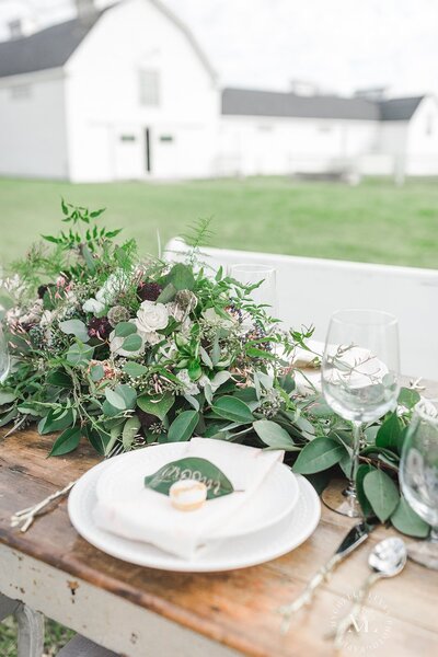 A beautiful table scape set up for a wedding. It has greenery on it as well as utensils and a white plate. In the background is a white barn