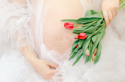 A pregnant woman holds her stomach and tulips, wearing a white tulle dress during a fine art photoshoot.