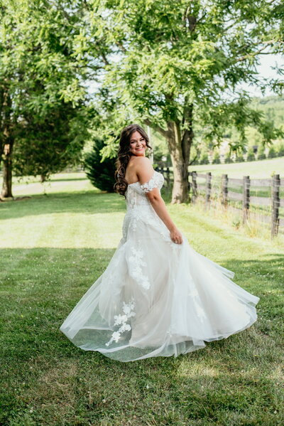 Bride in a flowing white gown twirling on a grassy path beside a historic barn wedding venue in Virginia, smiling joyfully under a clear sky. The dress features lace details and an off-shoulder design, perfectly complementing the rustic charm of Loudoun County's event spaces.
