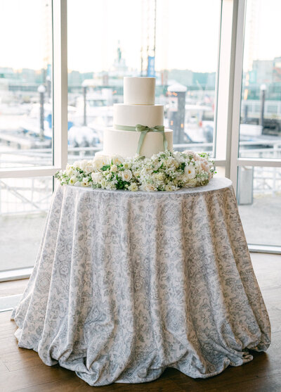 Three tier wedding cake on a round table covered in flowers at Current at Pier 59