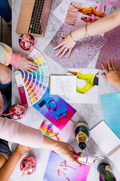 Top view branding photo of 5 women's hands sorting through a pantone book, glitter paper and inspiration photos