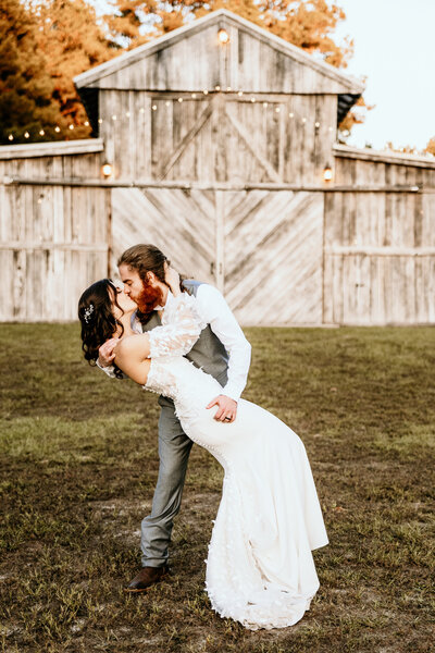 The groom dips his bride and kisses her outside the barn for their reception