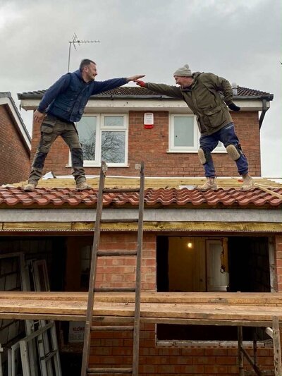 Two builders reaching toward each other while standing on a tiled roof.