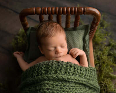 Newborn baby boy sleeping in a green knit wrap on a wooden bed with greenery, photographed by Austin newborn photographer Hello Photography