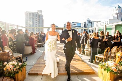 Bride and groom smile as they walk down the aisle with a city scape behind them