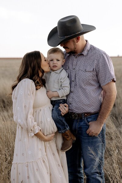 pregnant mom holding her two year old son with one hand kissing him on the cheek with dad standing next to them smiling down on both of them.