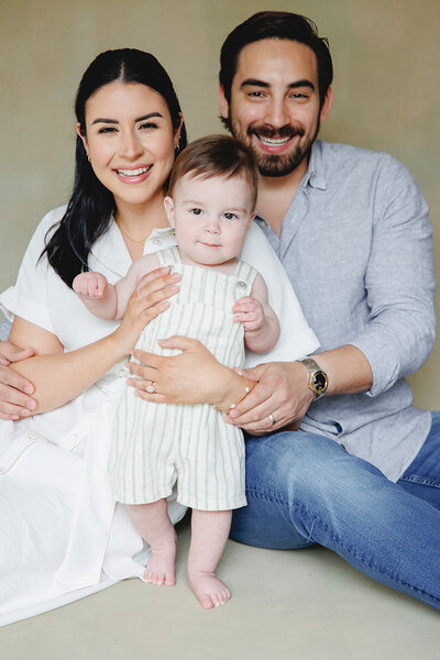 High end family portrait of mother and father with their young child all posed in front of a taupe backdrop