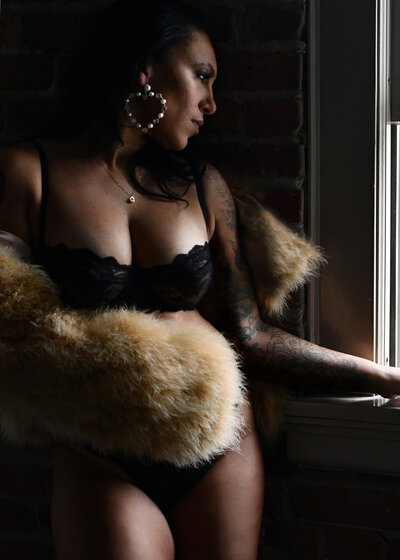 Woman looking through a window while wearing fur in her sexy boudoir photoshoot.