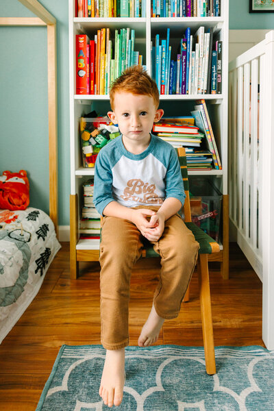 red headed young boy sitting on chair in front of bookshelf with books arranged in rainbow colors
