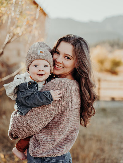 A young mother looks over her shoulder, smiling as she holds her six month old baby boy who is adorned with a Carhartt beanie and denim jacket. The setting is a mountainside in Colorado.