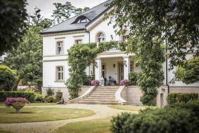 The front of a large, white estate with a looping driveway and climbing vines across the house,