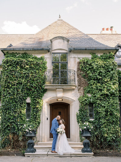 A bride tilts her face up for a kiss from her groom as they stand on the porch of a manor house covered with ivy and surrounded with flowers. | Wedding Photographer in Pittsburgh | Anna Laero Photography