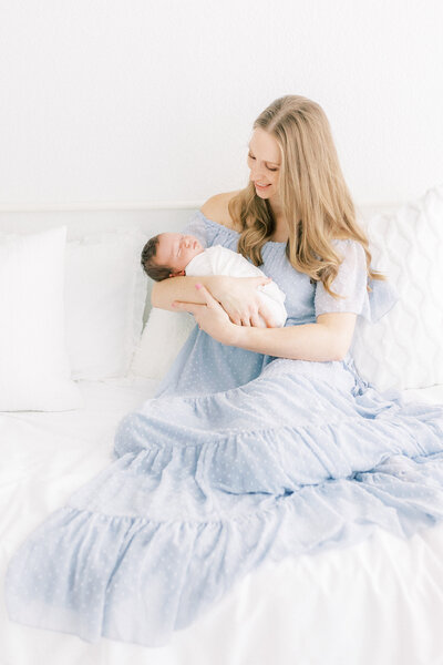 new mom holding baby boy in a blue dress sitting on bed in white studio