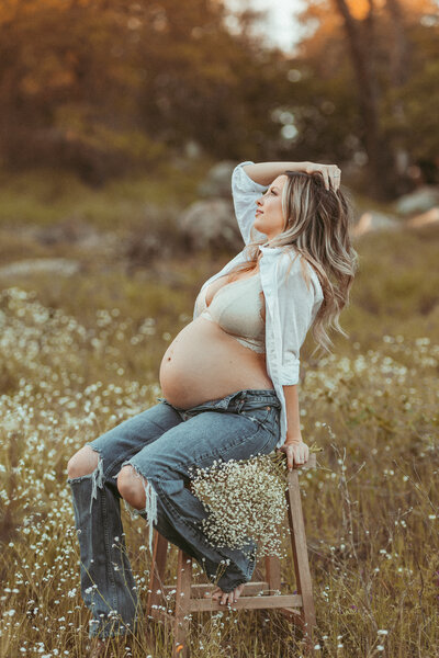 A pregnant mom in jeans and an unbuttoned white shirt poses on a stool in a wildflower field at sunset. 