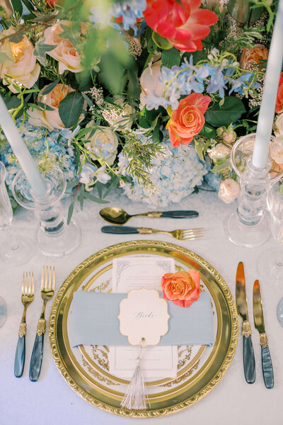 wedding table setting showing a layered gold charger plate wrapped in a blue napkin topped with a natural paper menu and coral flower