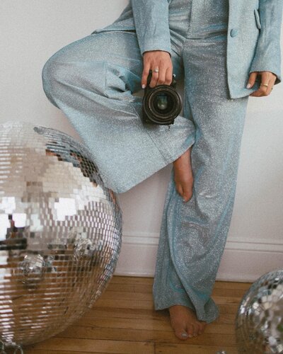 A woman leaning against a wall in a blue suit holding a camera next to a large disco ball