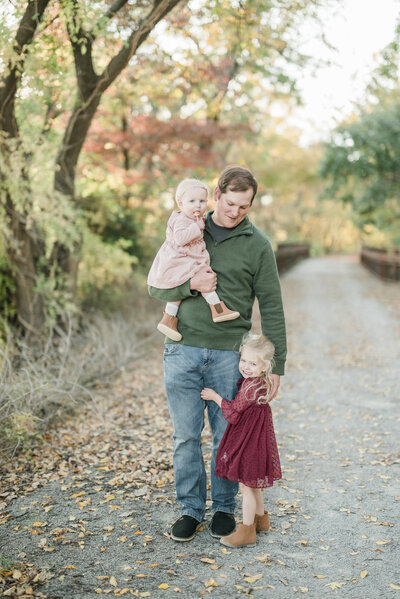 Father holding a baby girl and walking with his older daughter on a tree-lined path during a fall family session with a Kansas City photographer.