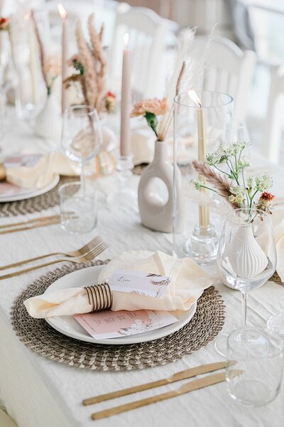 Soft and romantic wedding tablescape with woven placemats, blush menus, ivory napkins, and minimalist ceramic vases with dried florals