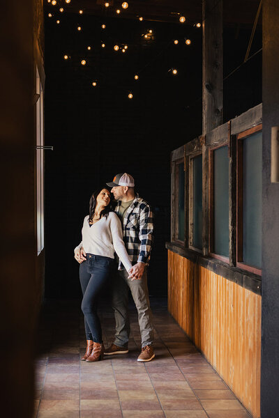 A couple stands in a warmly lit hallway, captured by Brainerd Lakes Area Photographer. The man, in a plaid shirt and cap, embraces the woman in a white sweater and jeans. They both smile beneath the string lights above, their happiness perfectly framed by Amber Bauer Photography.