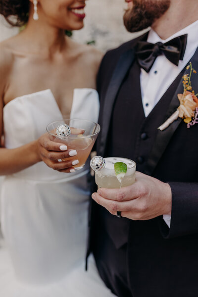 bride and groom smile at each other while holding cocktails with disco ball stirrers