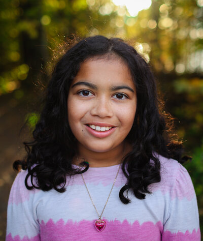 A smiling girl with curly hair, wearing a pink tie-dye shirt and heart necklace, outdoors with sunlight filtering through the trees at Lemoine Point.