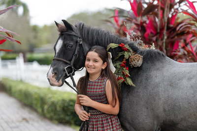 a blue roan pony with a wreath on its neck being held by a young girl in front of a plant with red leaves