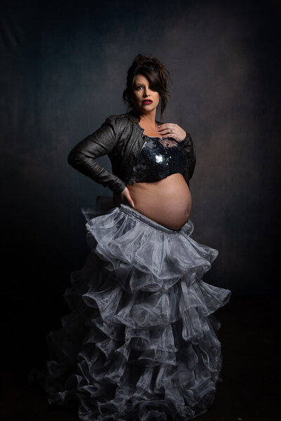Charlotte creative Maternity Photograph created in our Fort Mill studio on a molten fine art backdrop Mom looks dolled up and surprised by her situation wearing a sequin top, leather motorcycle jacket and a tulle fluff skirt with her belly exposed
