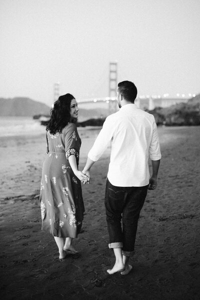 Classic black and white engagement photography at Baker Beach, San Francisco. Timeless candid moment with couple walking barefoot along the shore, featuring the iconic Golden Gate Bridge in the background. Floral midi dress and casual white shirt styling. Fine art film photography capturing authentic San Francisco landmarks.