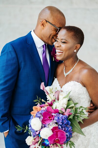 Groom wears a navy blue suit and smiles at bride holding a bright bouquet of flowers.