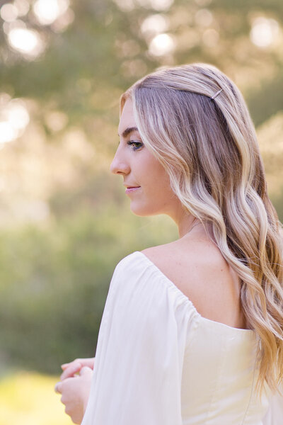 A close-up of a young woman with long, blonde hair wearing a white, long sleeve blouse, looking to her left with her back facing the camera in San Juan Capistrano.