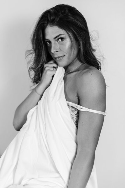 A black and white image of a women with her bra strap pulled downed covered by a sheet. 
