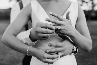 Groom standing behind Bride with their hands in front of Bride putting on wedding bands