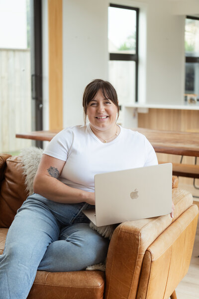 Chantal Hewitt smiling holding her laptop while wearing a white shirt and sitting on a leather tan touch - Family Autism Support Coach at Chantal Hewitt Coaching10
