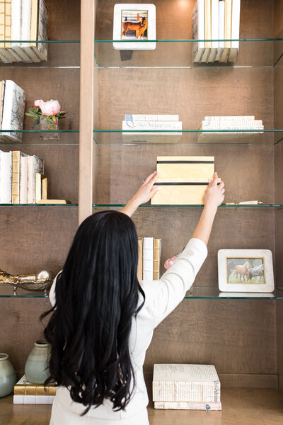 Kam is organizing a wooden portrait with both of her hands. She is standing in front of a closet with books and home decoration . Kam has black long hair and she is wearing a white t-shirt